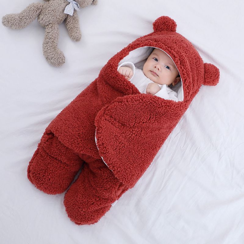 Baby in a red bear-shaped sleeping bag on a white surface with a teddy bear beside it.